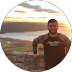 A bearded man leaning on a stone wall overlooking a scenic sunset and lake view.